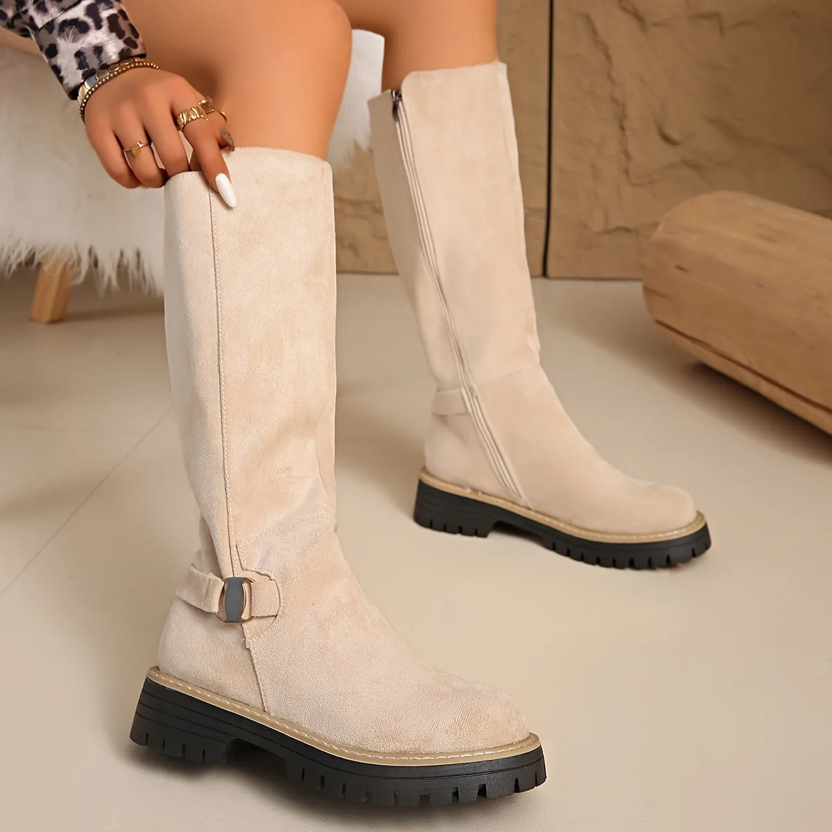 Close-up of beige suede knee-high boot with side zip and metal buckle detail, hand adjusting the shaft