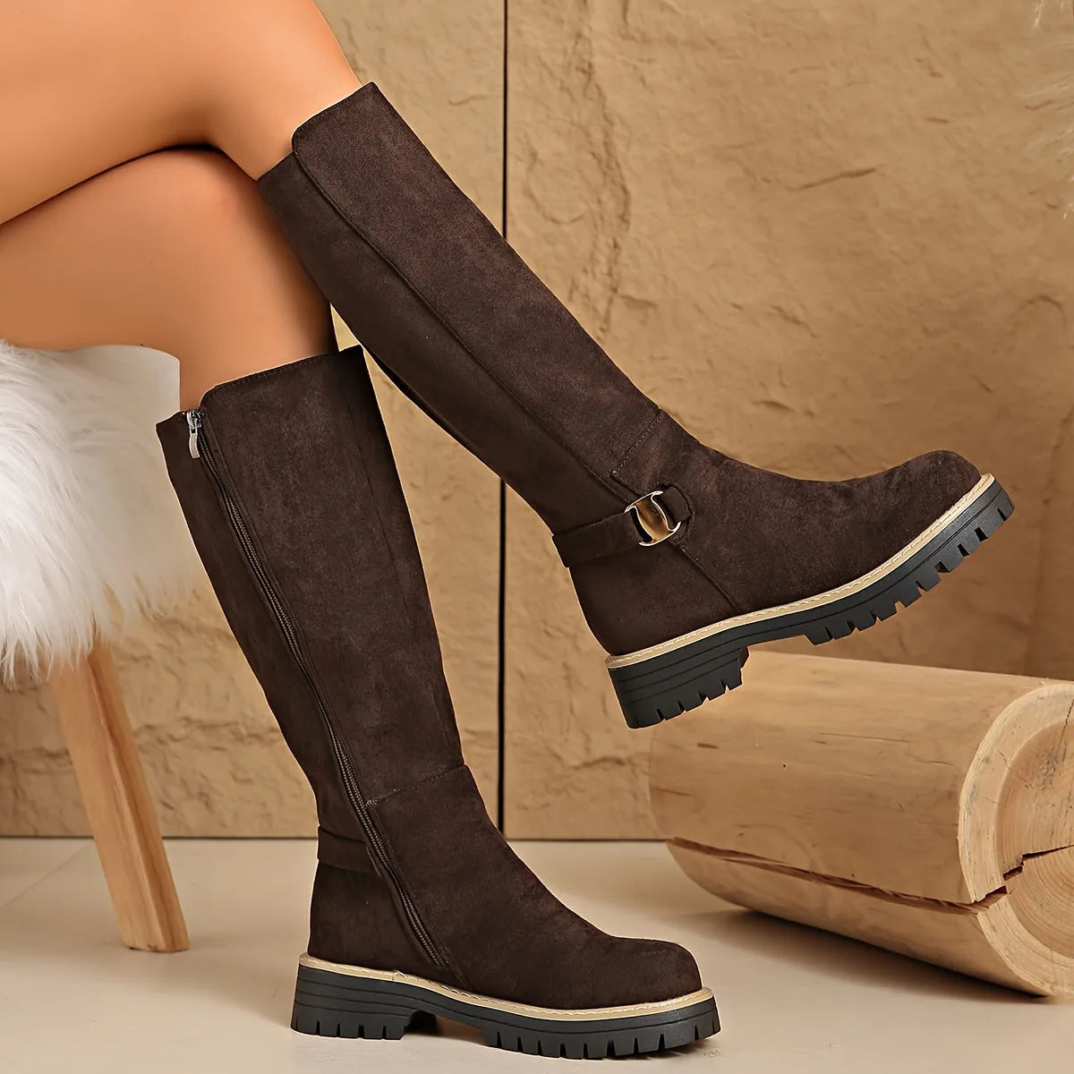 Pair of coffee brown suede knee-high boots with chunky ridged sole, one boot propped on a wooden log
