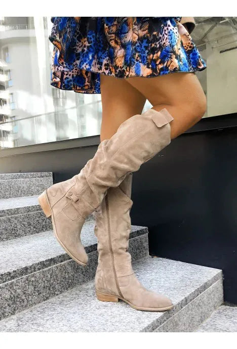 Beige suede knee-high slouch boots worn on stone steps, showing the ruched shaft and side buckle strap detail