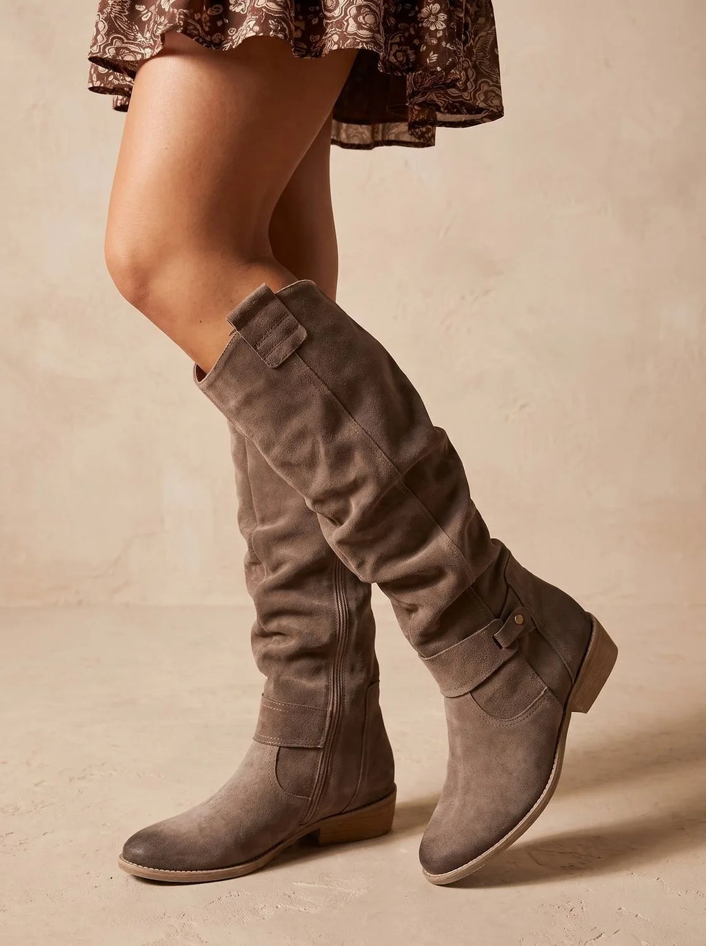 Taupe grey suede knee-high boots with ruched shaft and ankle strap, paired with a floral print mini dress