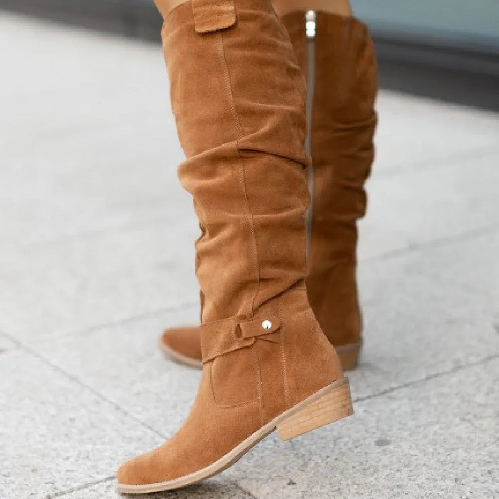 Tan suede knee-high boots shown close up, focusing on the buckle strap, pull tab and rounded toe