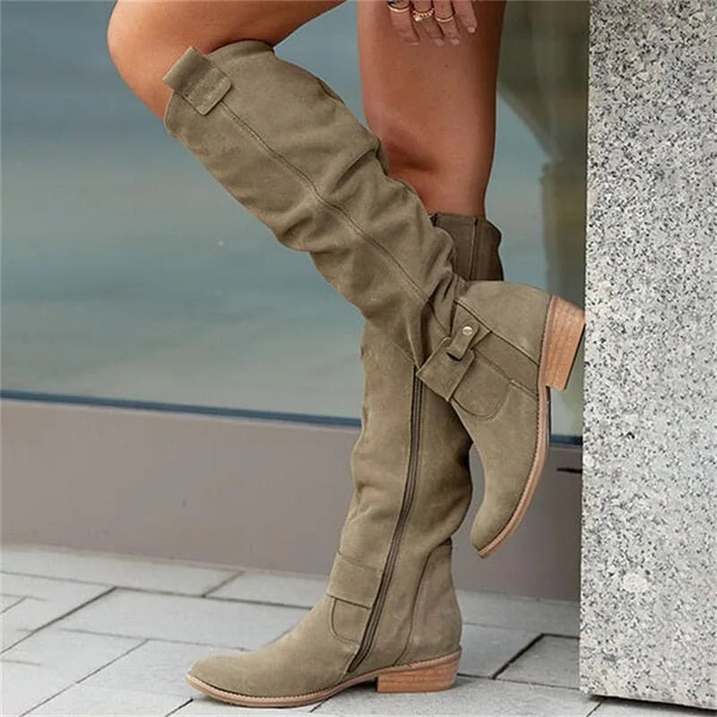 Khaki green suede knee-high boots against a wall, showing the pull tab, buckle strap and side zip