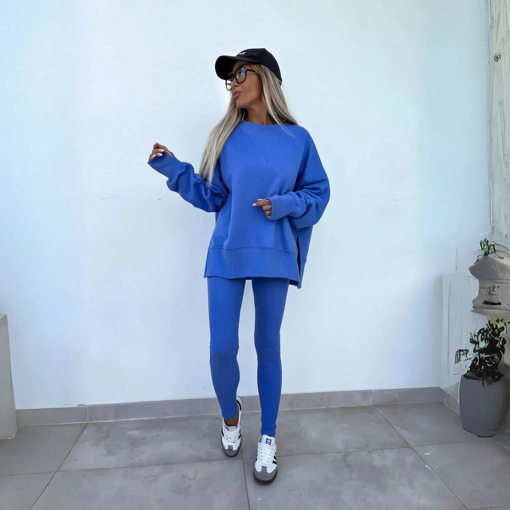 Model wearing a blue oversized crewneck sweatshirt and matching leggings against a white wall, paired with trainers