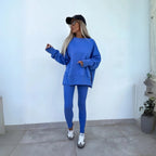 Model wearing a blue oversized crewneck sweatshirt and matching leggings against a white wall, paired with trainers