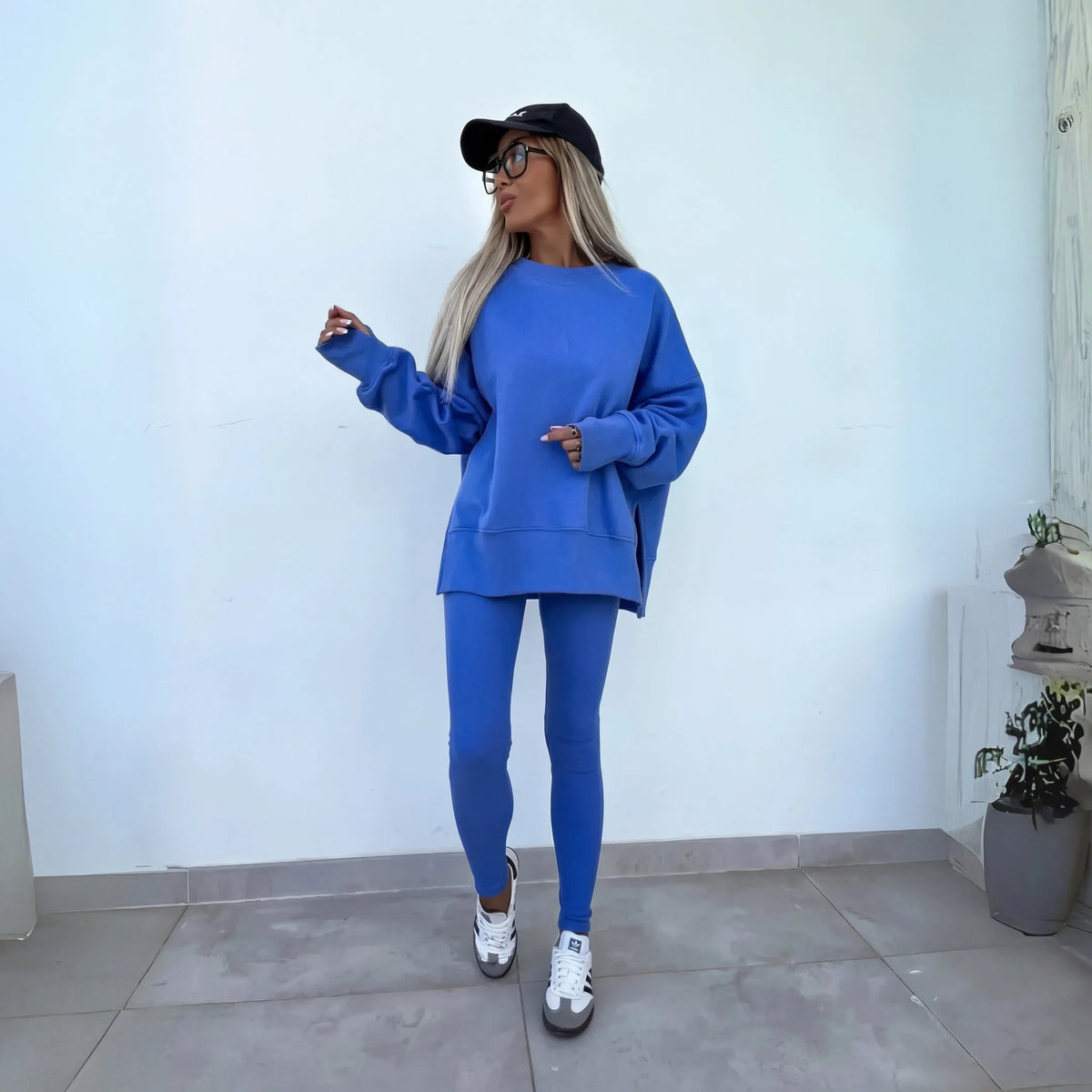 Model wearing a blue oversized crewneck sweatshirt and matching leggings against a white wall, paired with trainers