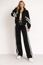 Full-length view of a black zip-up tracksuit jacket with white sleeve stripes worn with white-striped wide-leg joggers and trainers
