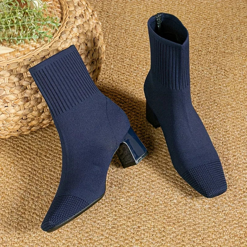 Navy knitted sock boots with square toe and block heel, paired flat on a jute rug beside a woven basket