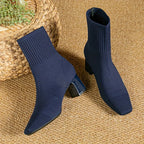 Navy knitted sock boots with square toe and block heel, paired flat on a jute rug beside a woven basket