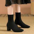Model wearing black knitted sock boots with square toe, standing on a woven rug under a sheer black skirt