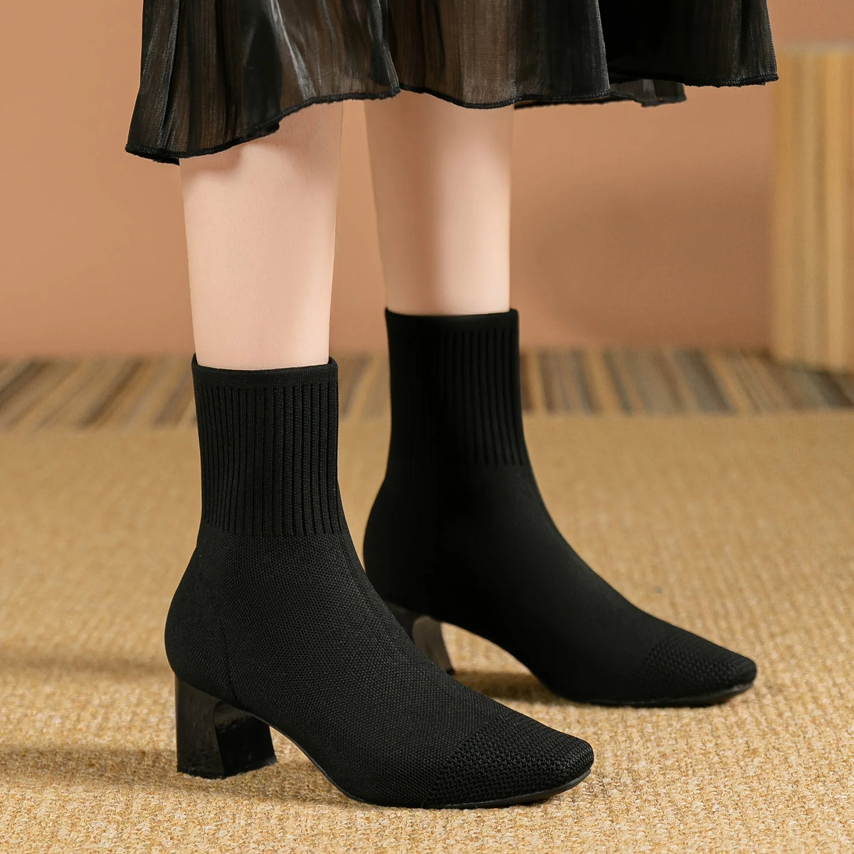 Model wearing black knitted sock boots with square toe, standing on a woven rug under a sheer black skirt