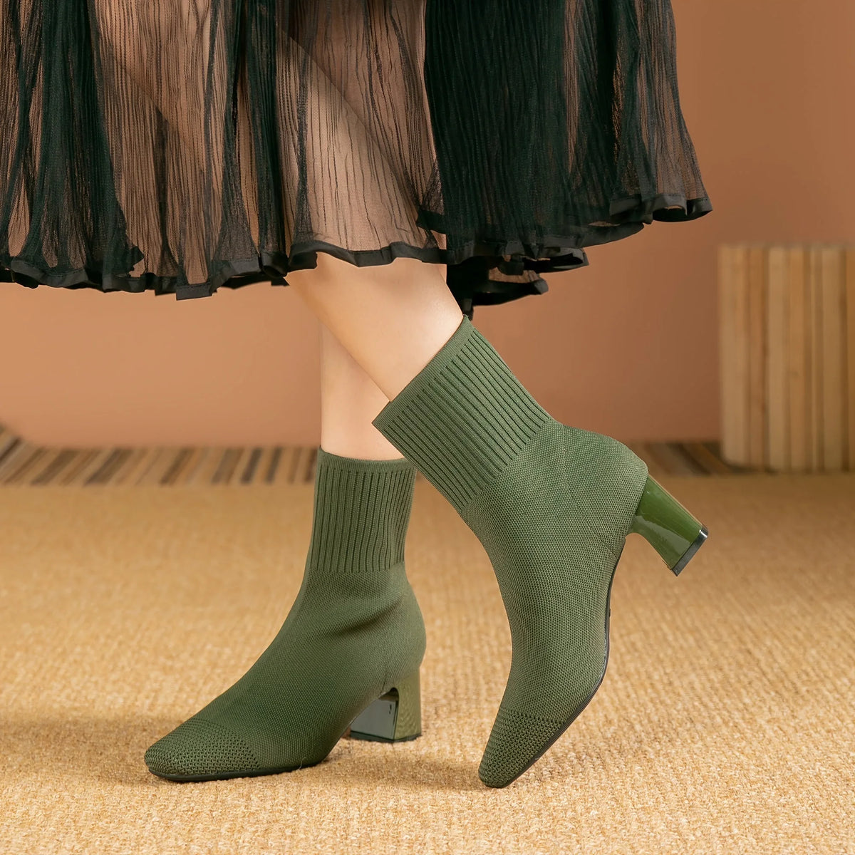 Close-up of model's legs in olive green sock boots, one foot lifted to show the block heel profile
