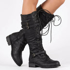 Model wearing black lace-up knee-high boots with buckle straps, side zip and chunky sole, crossed legs