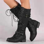 Close-up of black lace-up knee-high boots with buckle straps, worn on legs against a plain grey background.