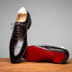 Side profile of two dark brown brogue oxford shoes on a wooden surface, one resting on its red sole with shoe trees inside