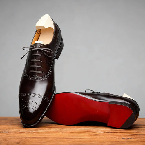 Side profile of two dark brown brogue oxford shoes on a wooden surface, one resting on its red sole with shoe trees inside