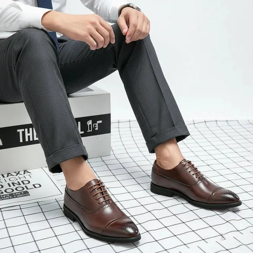 Man seated wearing brown leather cap-toe brogue oxford shoes with a suit and tie, side view against a patterned floor