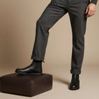 Man standing with one foot on a leather ottoman, showing black Chelsea boots with tailored trousers
