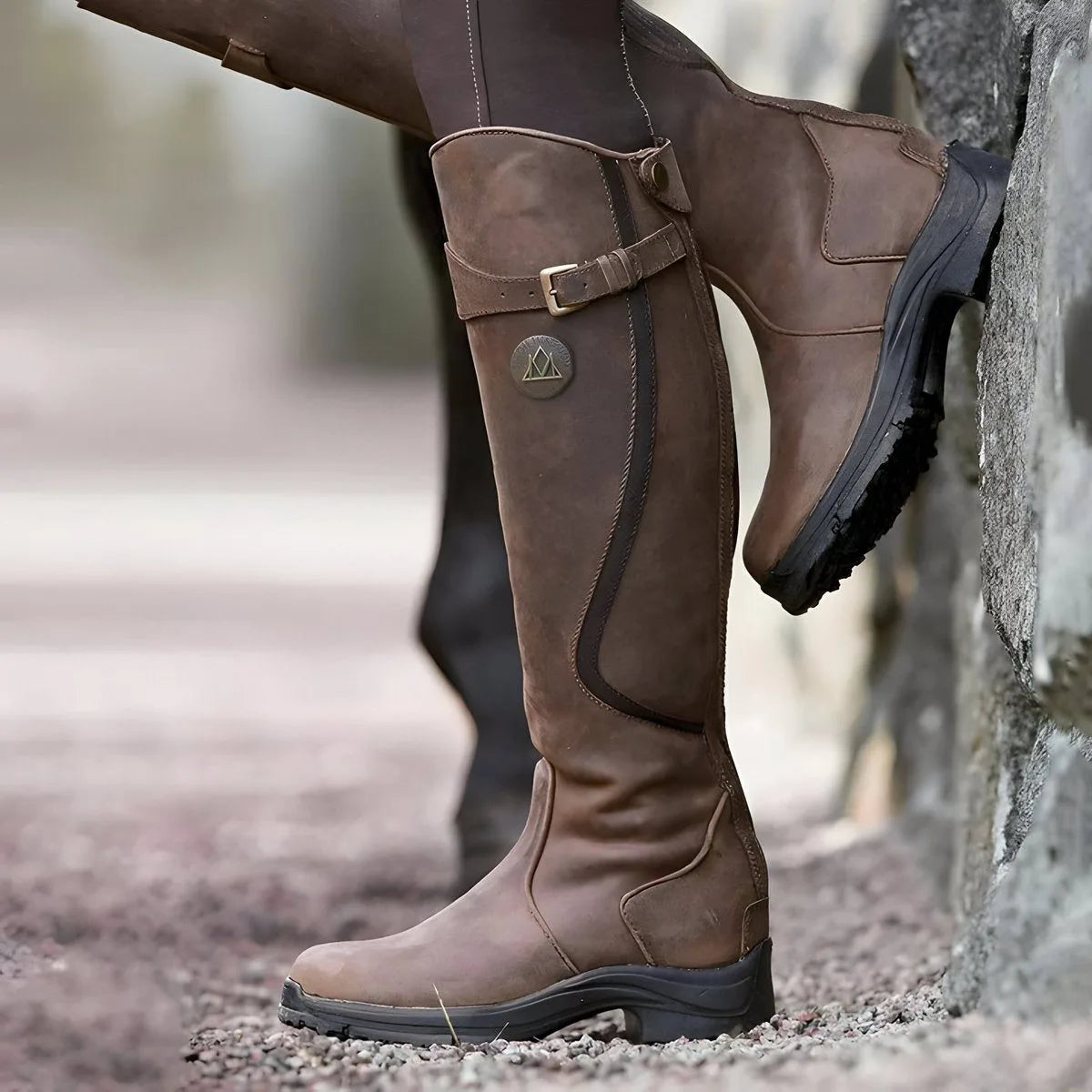 Side view of dark brown leather knee-high riding boots with buckle strap, worn while foot rests against a stone wall