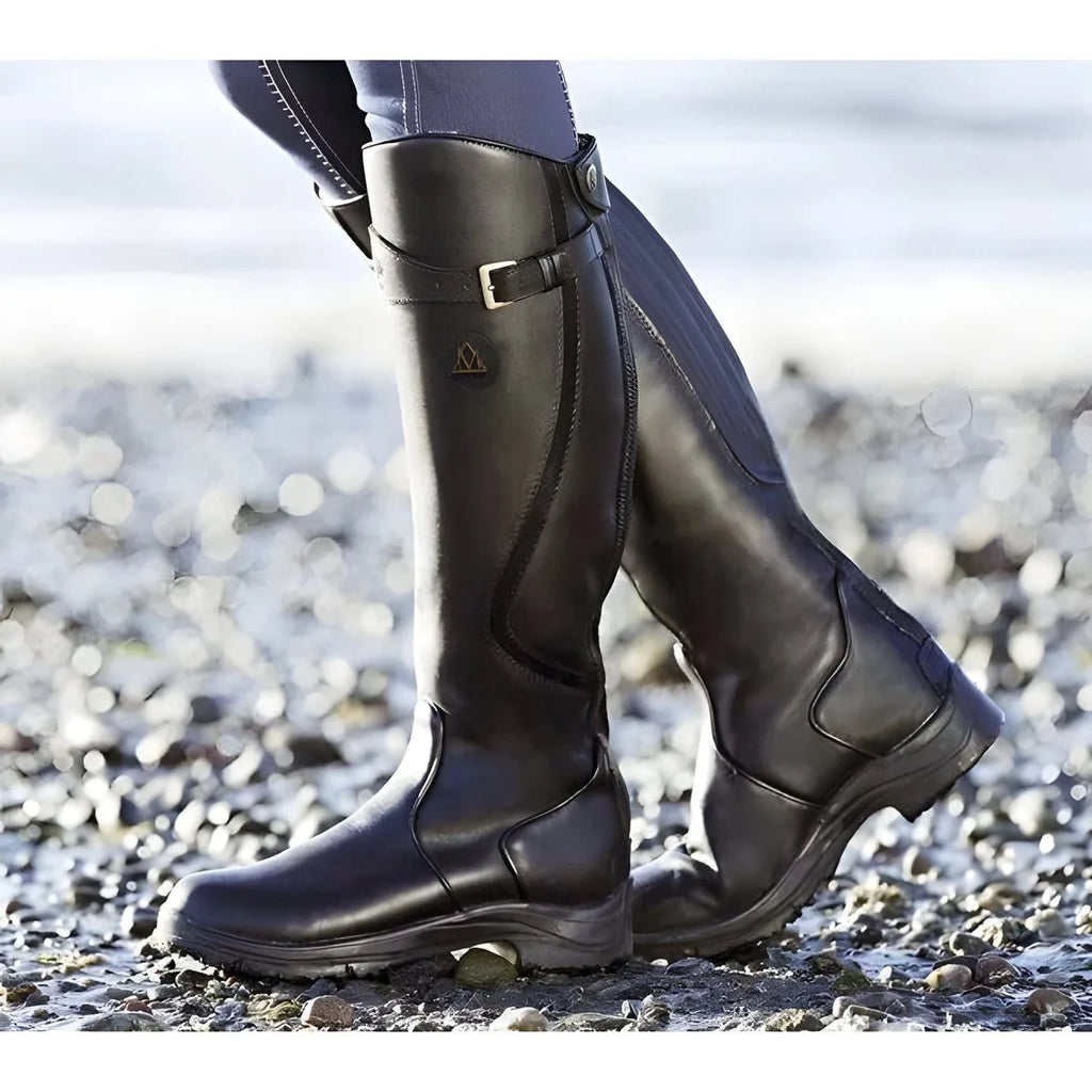 Sleek black leather knee-high riding boots with buckle strap and contrast piping, shown outdoors on pebbles