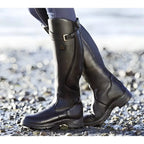 Sleek black leather knee-high riding boots with buckle strap and contrast piping, shown outdoors on pebbles