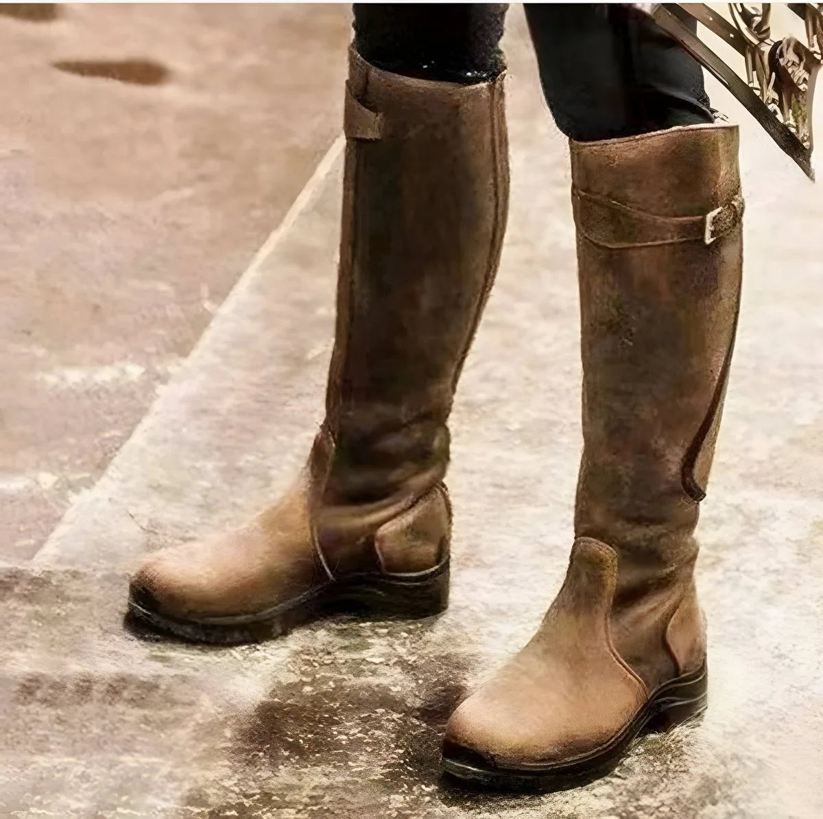 Studio-style shot of worn brown leather knee-high boots with buckle strap and pull tab, standing on stone ground