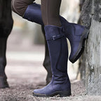 Navy blue suede-look knee-high boots with buckle strap, foot raised against a stone wall for detail