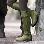 Olive green suede-look knee-high boots with buckle strap and shield emblem, foot propped against stone wall