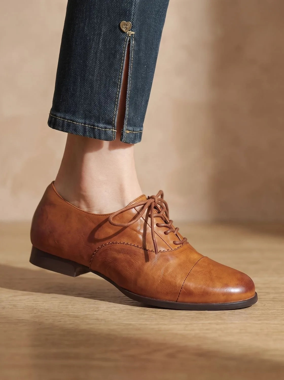 Side view of tan leather lace-up oxford shoe worn with cropped dark denim jeans, close-up on wooden floor