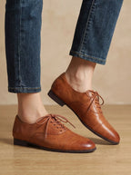 Side profile of both feet wearing tan leather oxford shoes with ankles crossed over dark denim hem