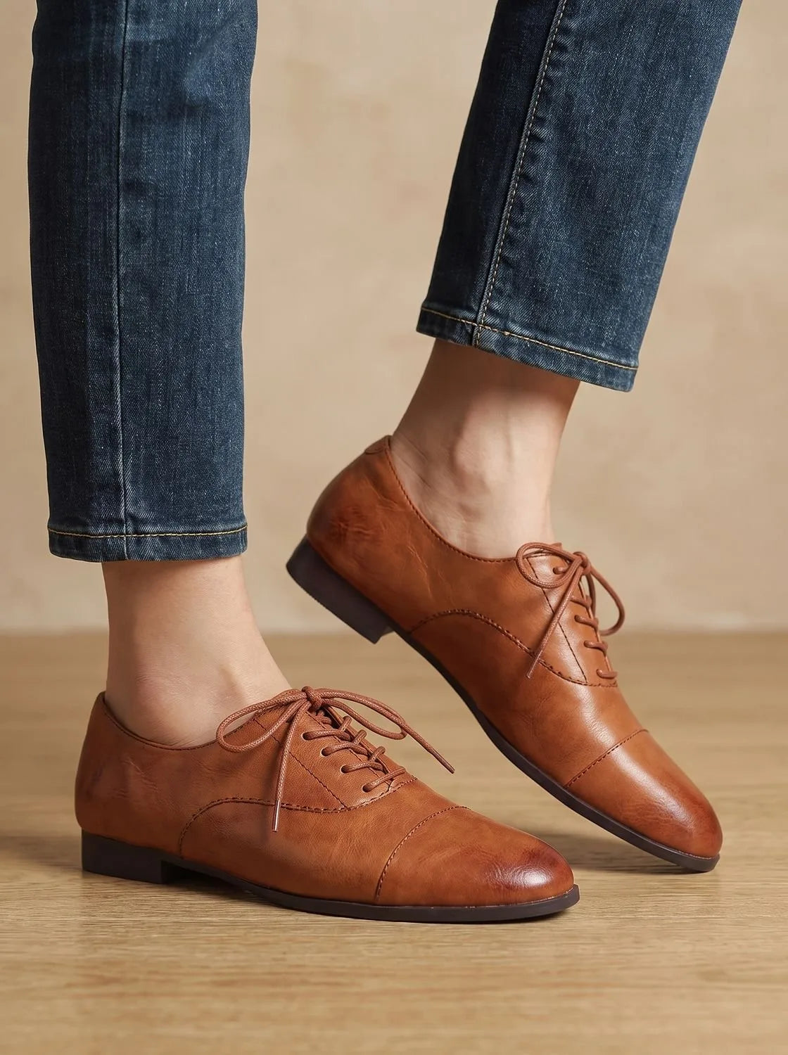 Side profile of both feet wearing tan leather oxford shoes with ankles crossed over dark denim hem