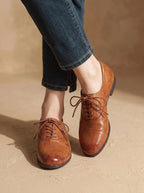 Two tan leather oxford shoes stacked artistically beside a dark driftwood branch on a beige textured surface