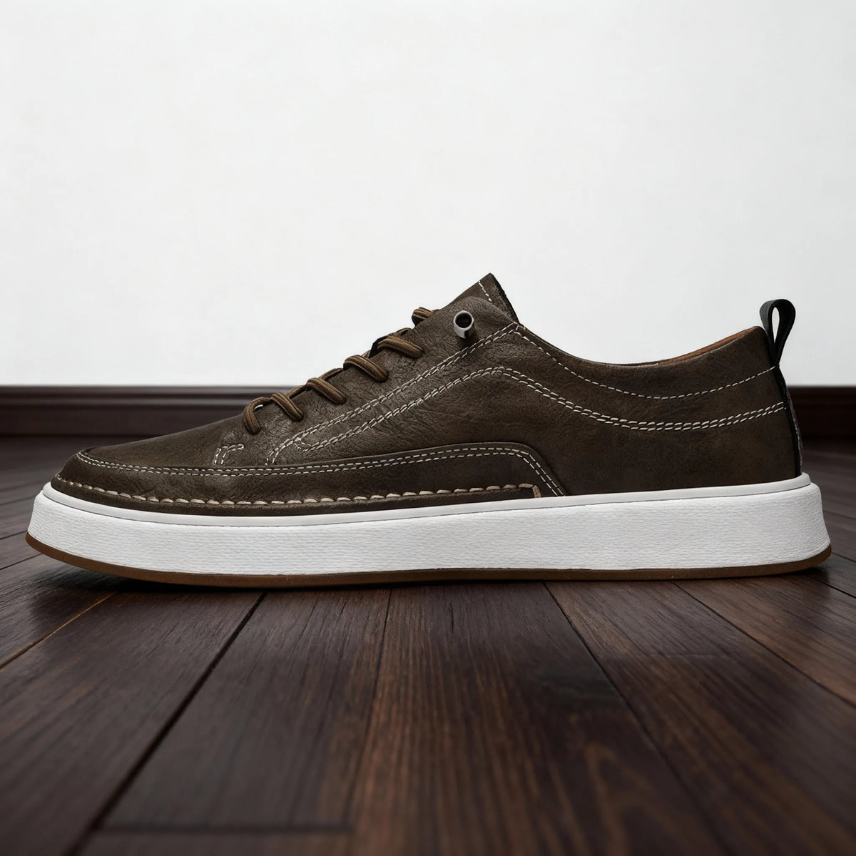 Full side profile of khaki brown leather trainer on wooden floor highlighting hand-stitched seams and pull tab
