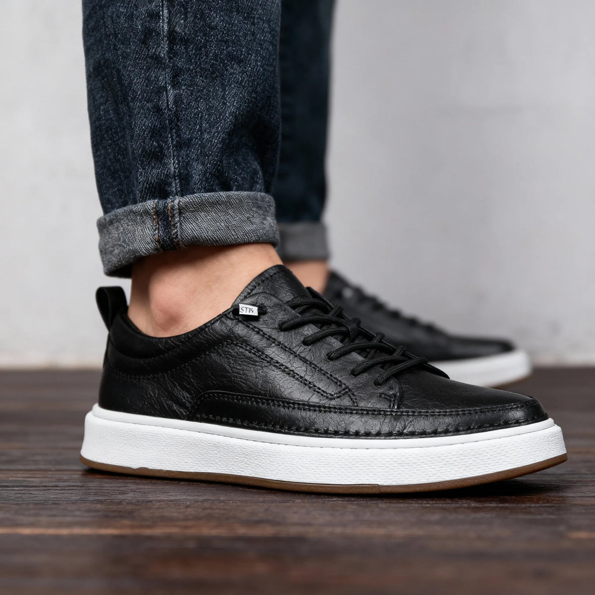 Close-up of black leather trainer worn on foot with rolled denim jeans, showing lace-up front and padded collar