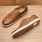 Pair of dark brown leather lace-up trainers with tan laces and heel patch resting on a wooden floor