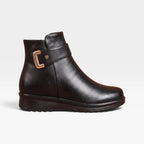 Side profile of black leather ankle boot with gold buckle strap and cushioned wedge sole against a plain backdrop
