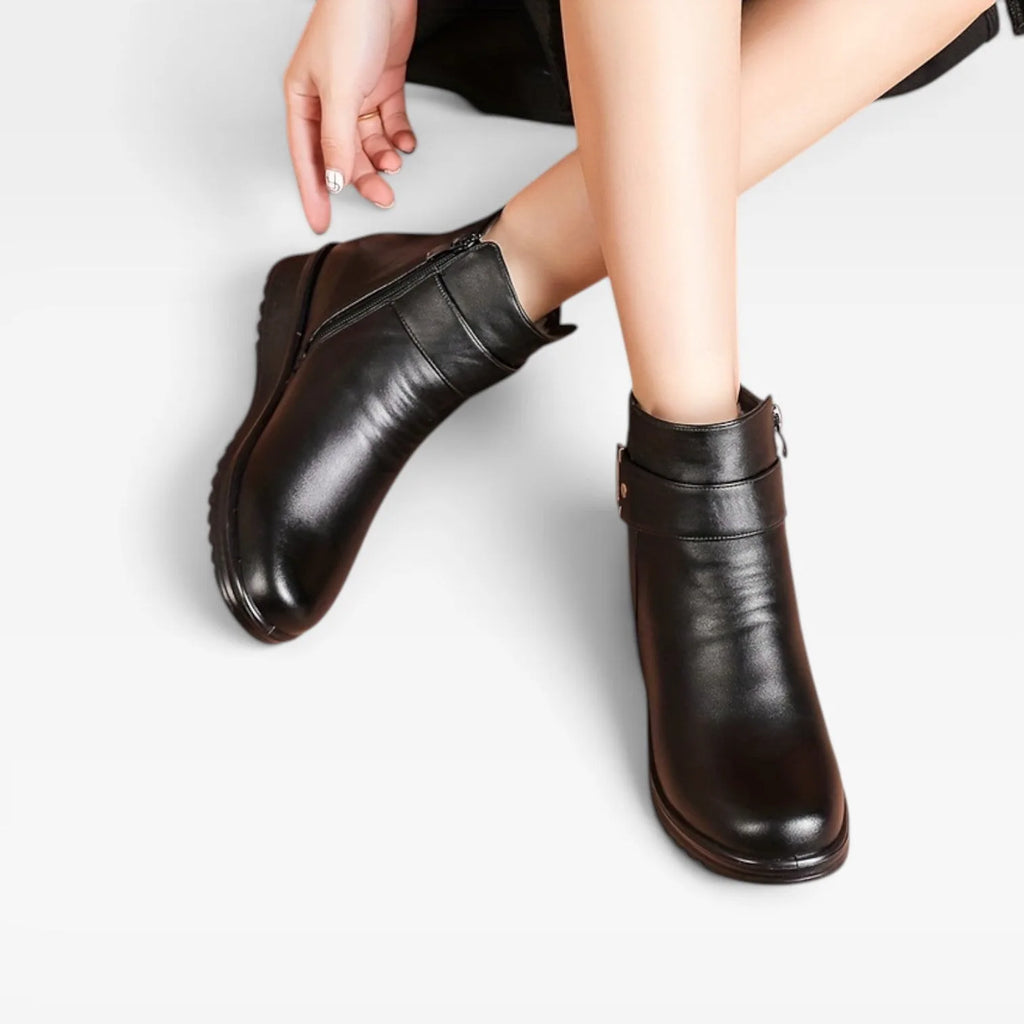 Close-up front view of black leather ankle boots with buckle strap and side zip, seated model with crossed ankles