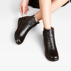 Close-up front view of black leather ankle boots with buckle strap and side zip, seated model with crossed ankles