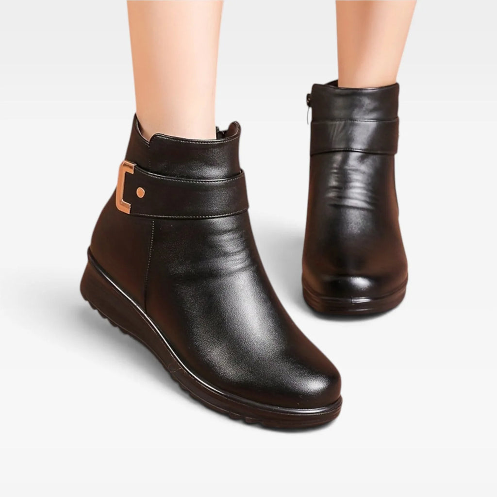 Front angled view of pair of black leather ankle boots on a model's feet, highlighting rounded toe and buckle trim