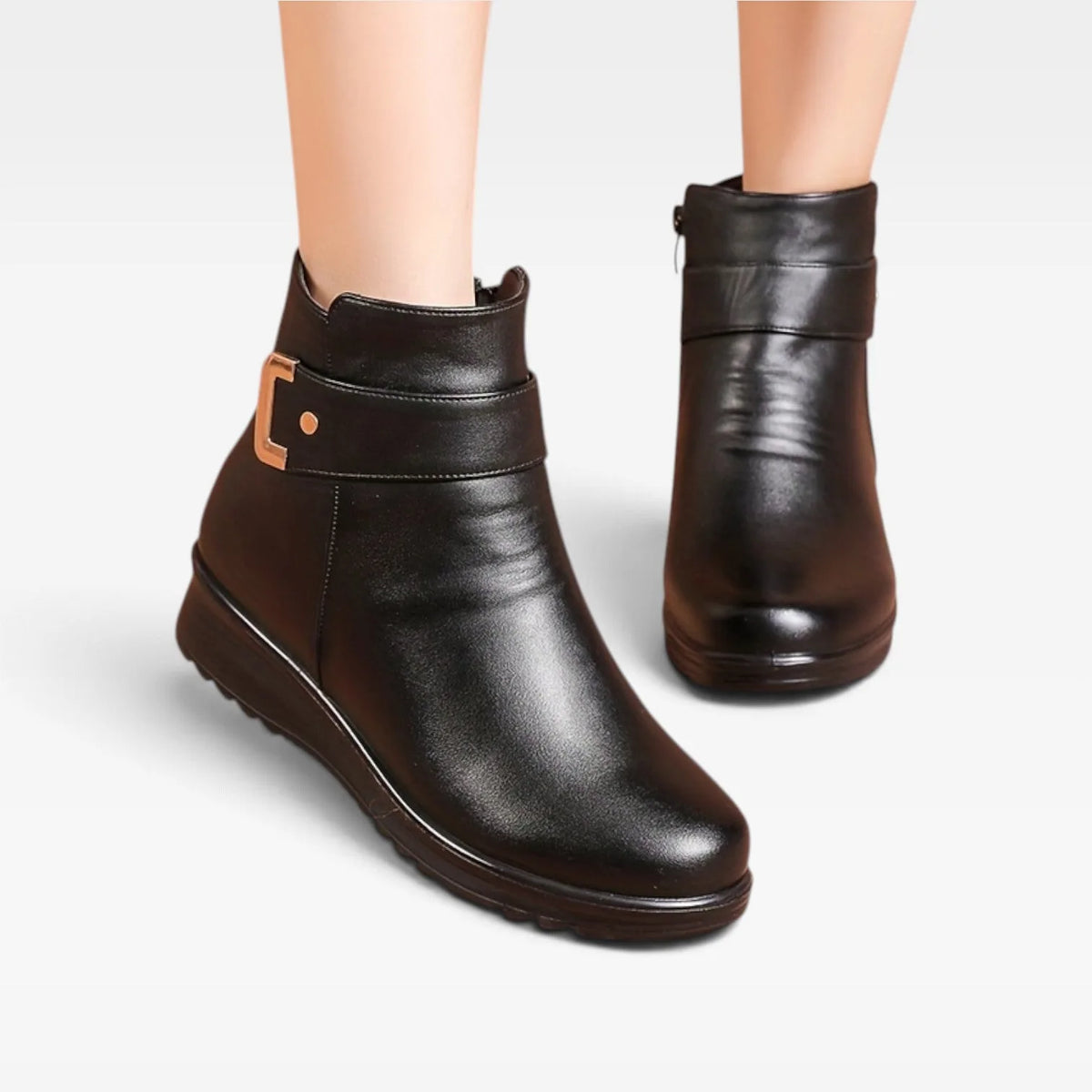 Front angled view of pair of black leather ankle boots on a model's feet, highlighting rounded toe and buckle trim