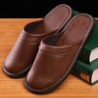 Pair of brown leather mule slippers with contrast stitching resting against green books on a wooden table
