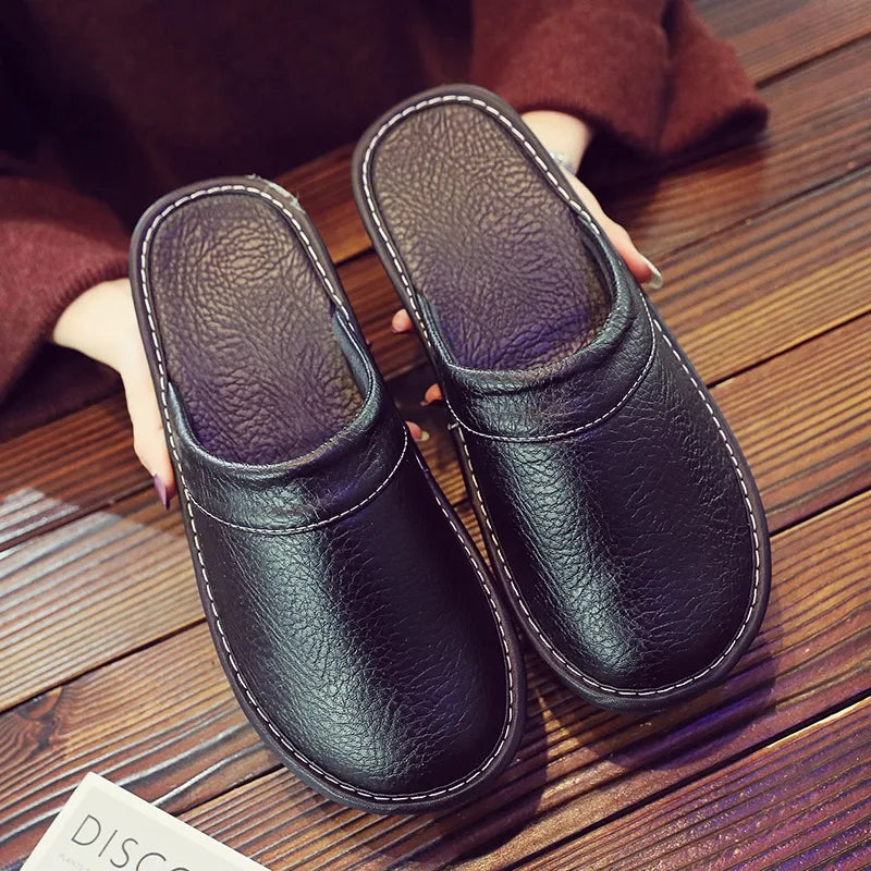 Overhead view of black leather slip-on slippers with white stitching held in hand on a wooden floor