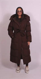 Front view of model in a long brown belted puffer coat with quilted panels, hood and tied waist belt