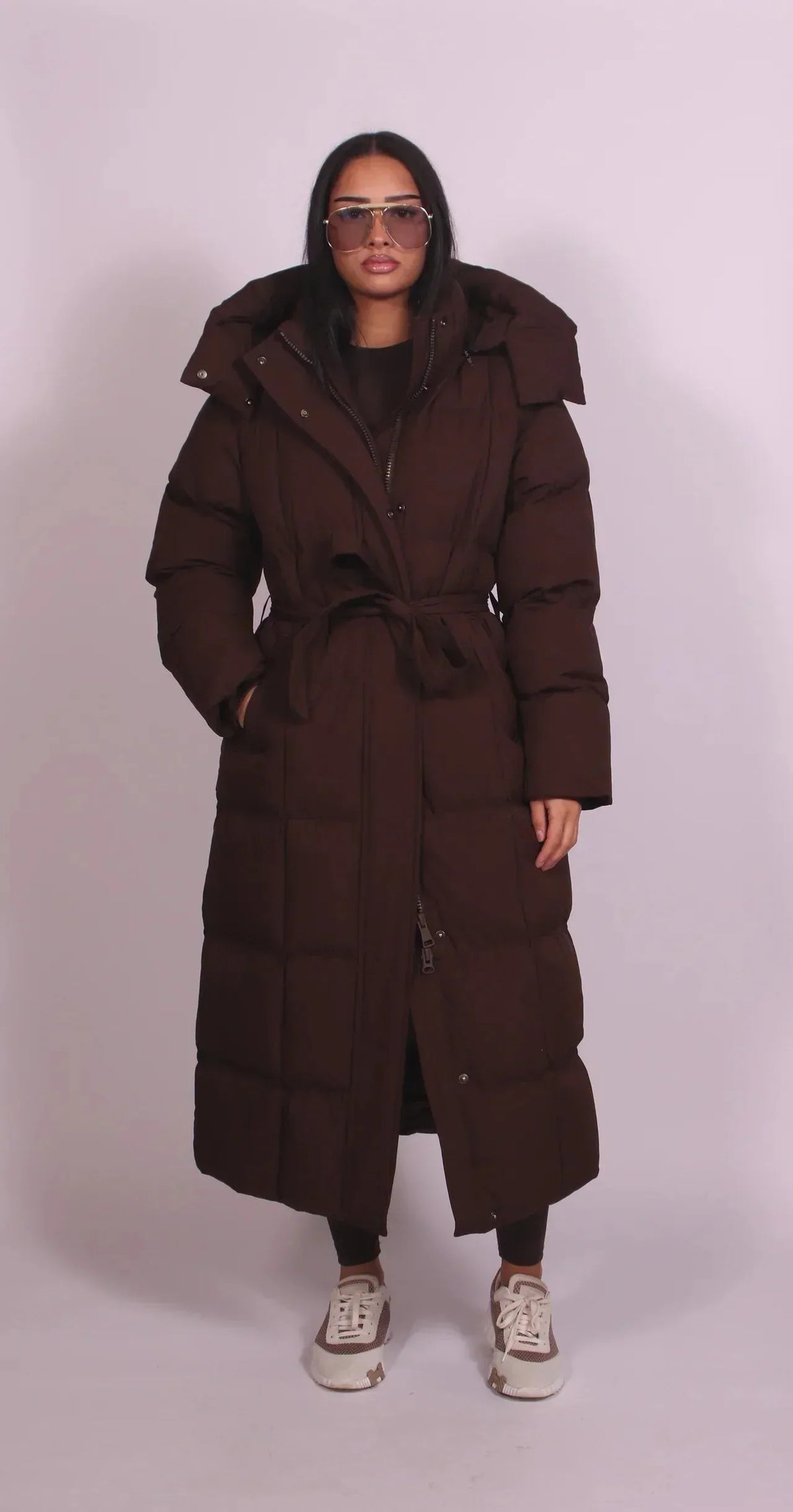 Front view of model in a long brown belted puffer coat with quilted panels, hood and tied waist belt