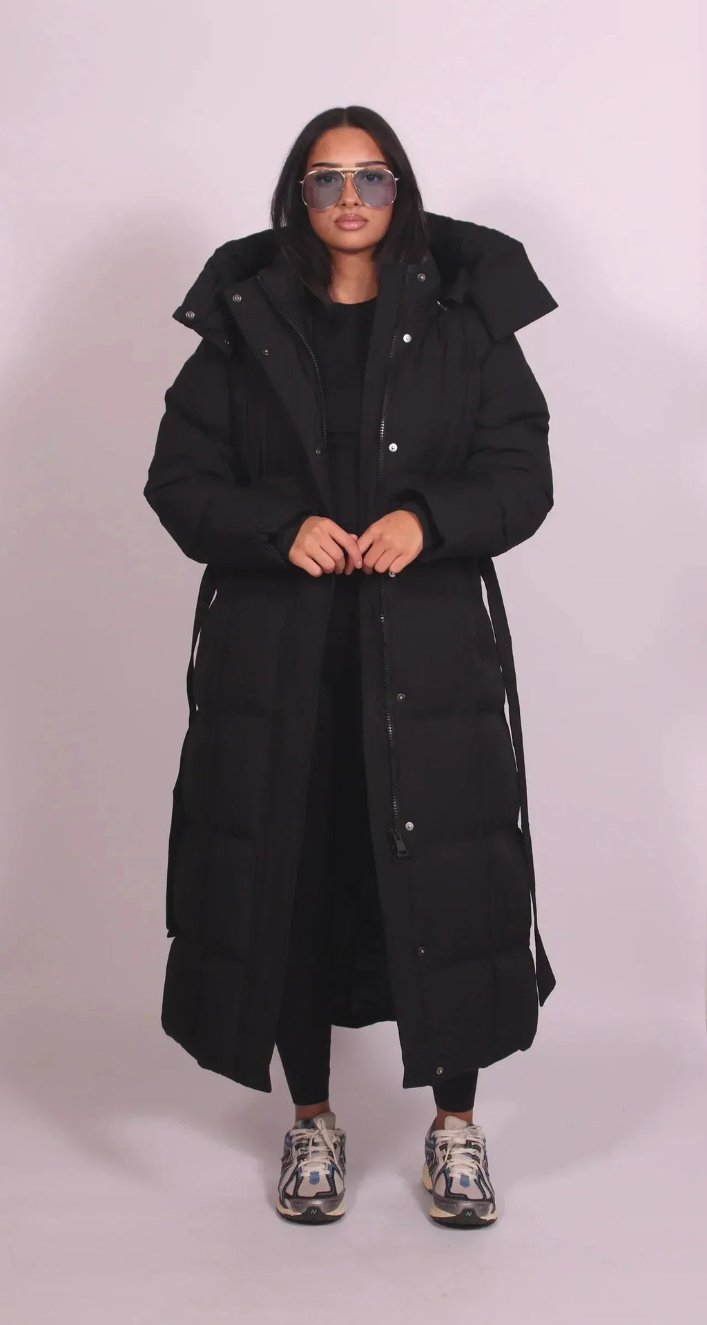 Front view of model wearing a long black belted puffer coat, zipped up with hood and side pockets
