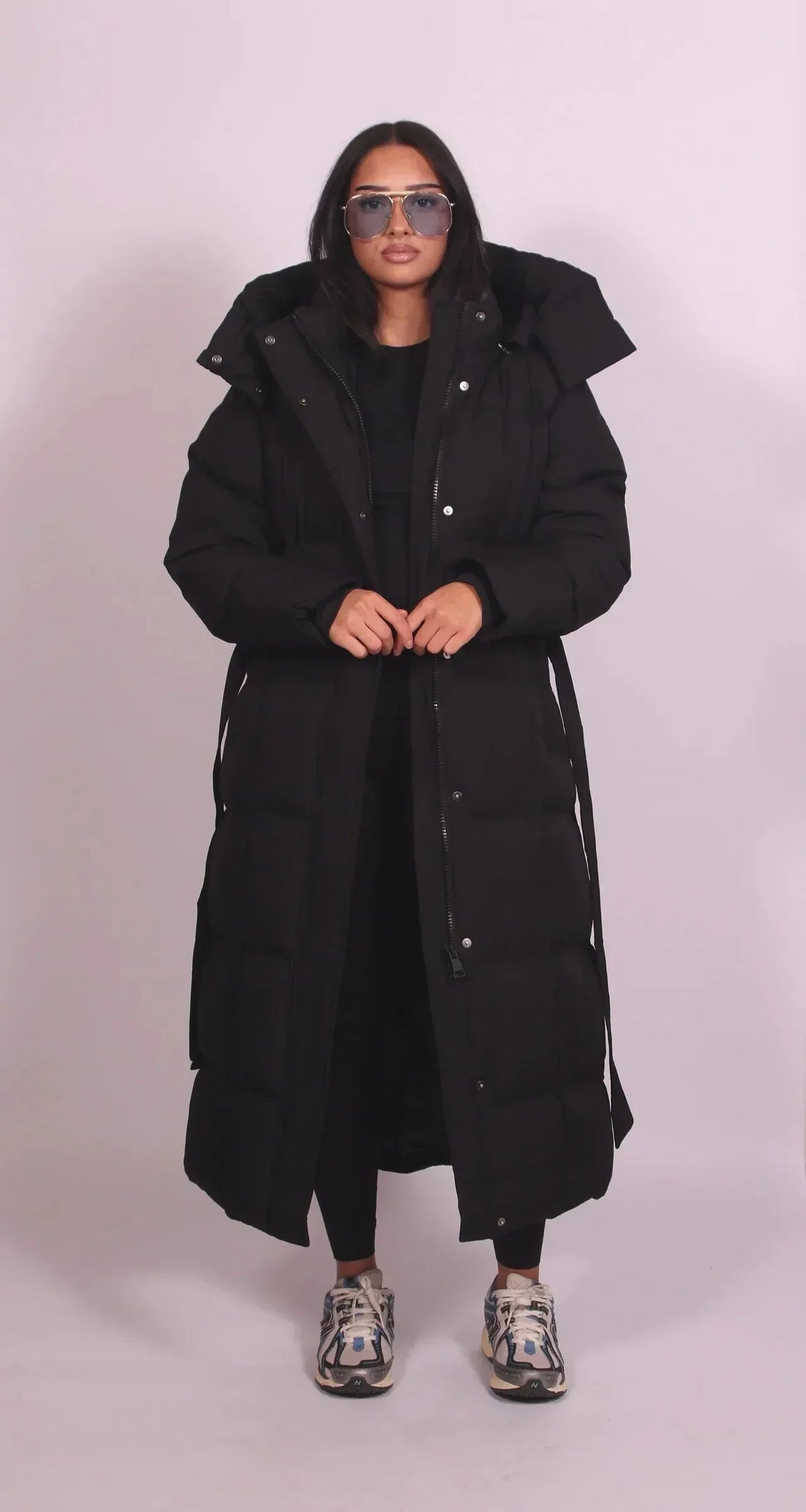 Front view of model wearing a long black belted puffer coat, zipped up with hood and side pockets