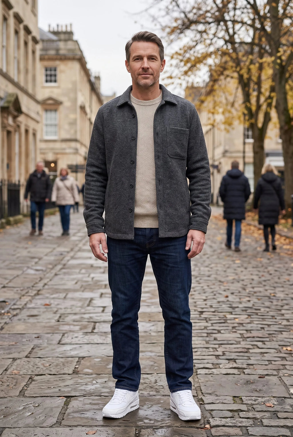 Man in grey wool overshirt, beige jumper and dark jeans wearing white trainers on a cobbled street