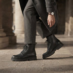 Close-up of a man striding in black leather combat boots on stone pavement, wearing a grey coat and wristwatch