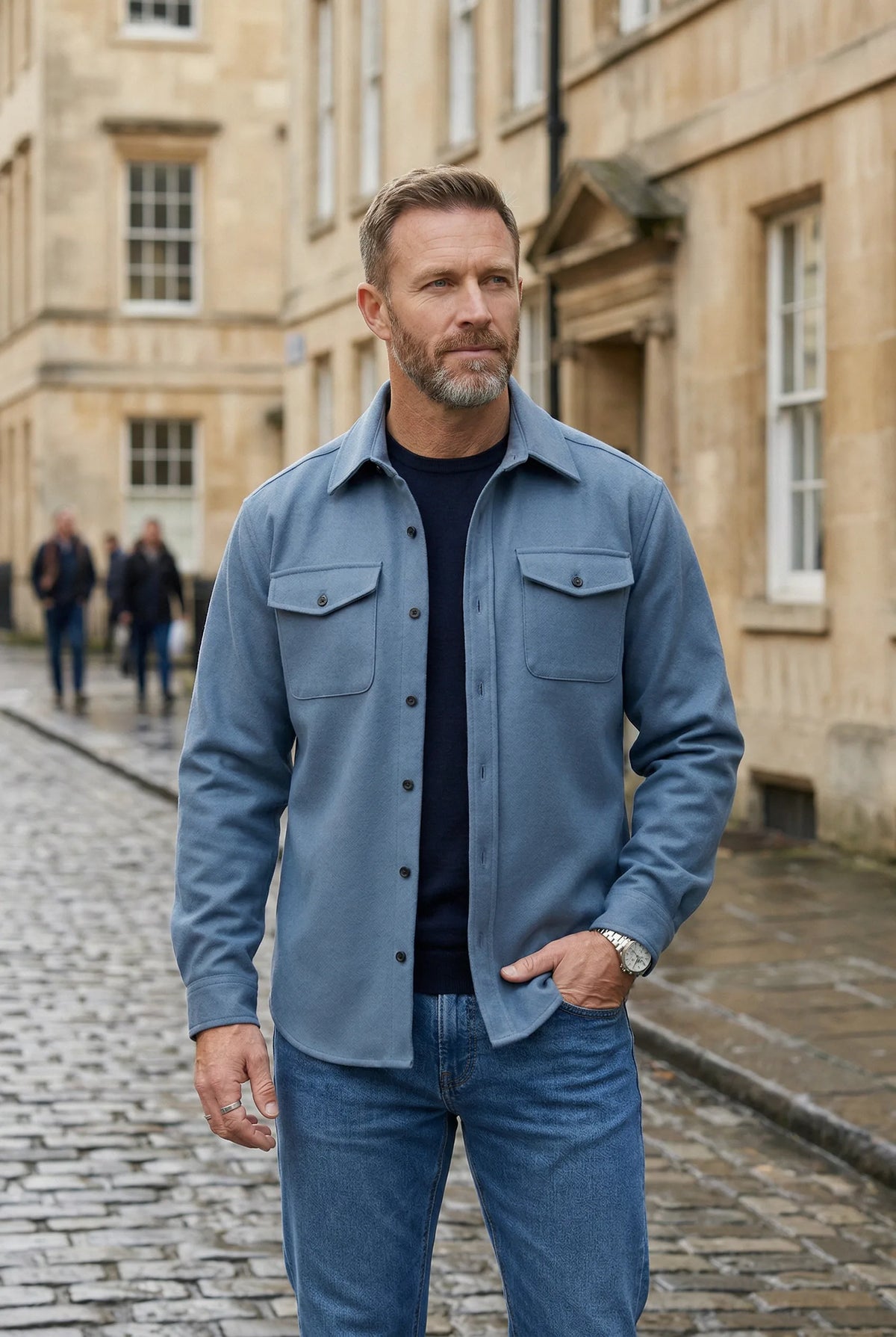 Man in a light blue overshirt and jeans walking along a cobbled street in a smart casual look