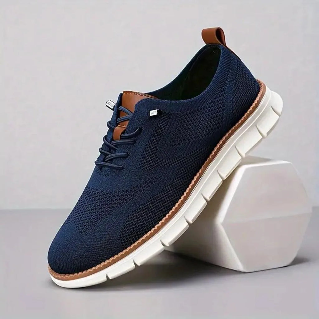 Navy blue knit sneaker with tan heel loop and lace eyelets resting against a white geometric block