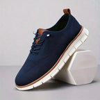 Navy blue knit sneaker with tan heel loop and lace eyelets resting against a white geometric block
